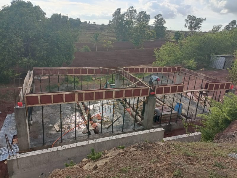 Nikita Textiles, Textile Park, Borgaon — site photograph of the reinforced concrete frame under construction, with red shuttering to the plinth beams, column starter bars and props in place, on farmland outside Borgaon.