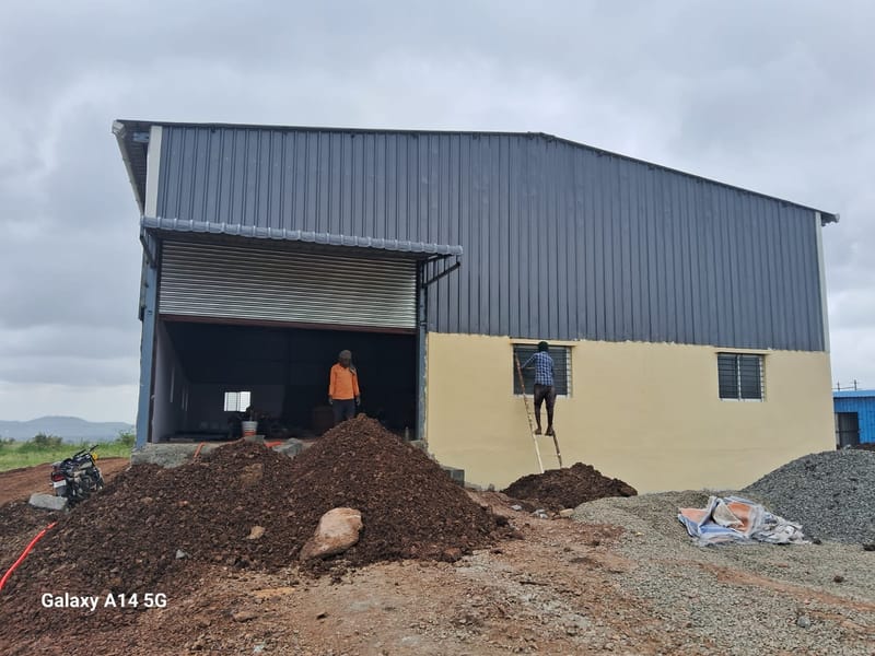 Textile industry shed for Shri Sai Textiles, Kanagala Industrial Area, KIADB — site photograph of the completed shed from the gable end, with dark grey profiled metal cladding, a cream plinth wall, a roller shutter opening and workers finishing the window openings.